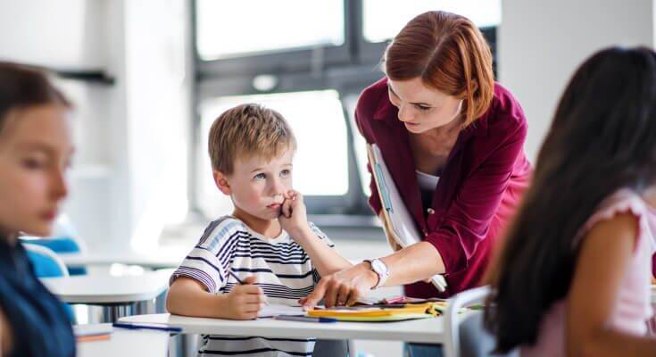 A female teacher leans over an elementary school student boy's desk and points at the paper to help him focus