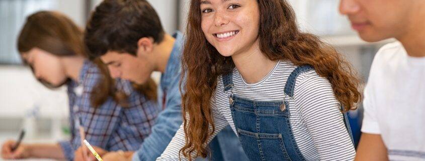 Satisfied young woman looking at camera while her friends studying at college. Team of multiethnic students preparing for university exam. Portrait of beautiful girl with freckles sitting in a row with her classmates during high school exam.