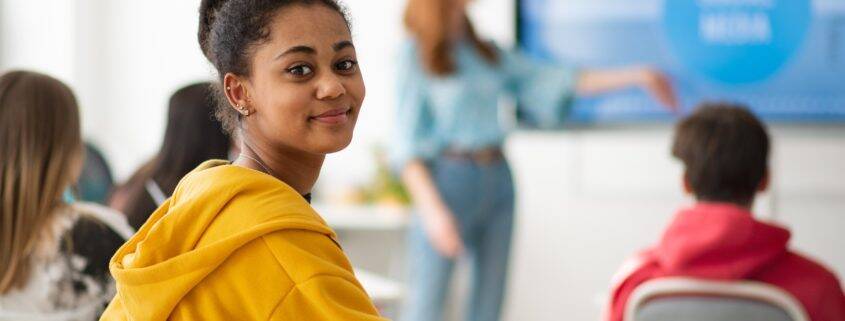 Happy college student sitting in school-desk and looking in camera.