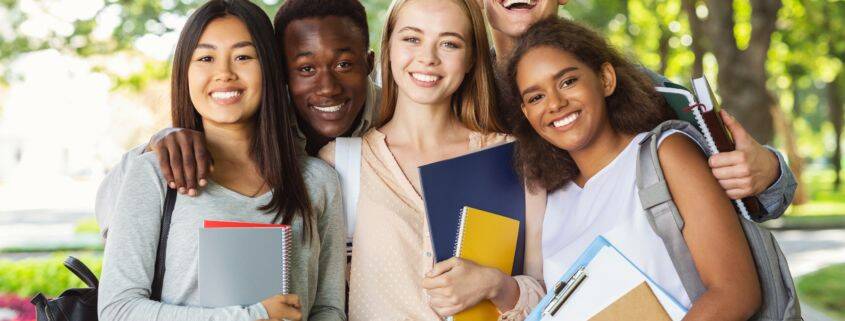 Group of international happy students with books and notebooks having fun in park after studying, smiling at camera