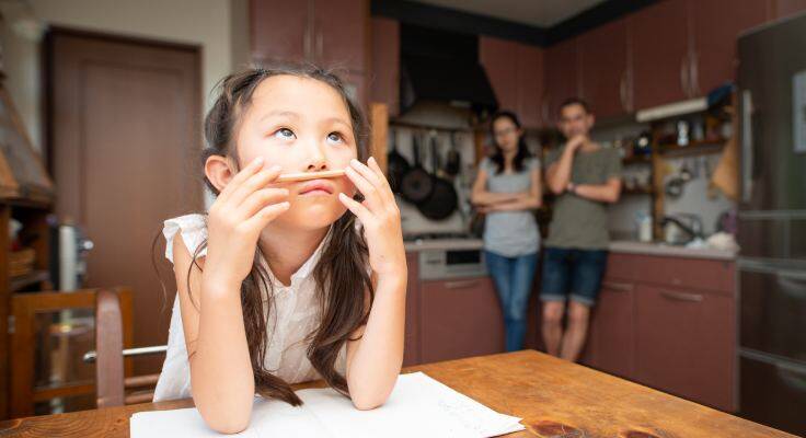 A bored little girl struggles with her homework as her concerned parents look on.