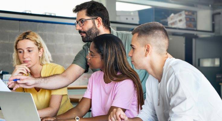 A bearded white male teacher actively instructing students using a computer monitor, facilitating a technology-enhanced learning experience.