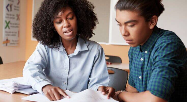 Female academic coach works with a middle school student. The coach reaches across a piece of paper to point at something, which the student looks at.