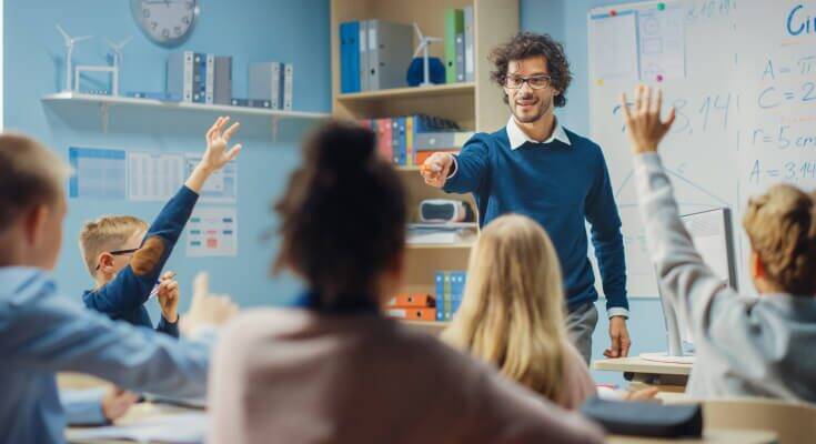 Male teacher in a blue sweater in glasses pointing at a student who is raising their hand. Teacher stands in front of a whtieboard.