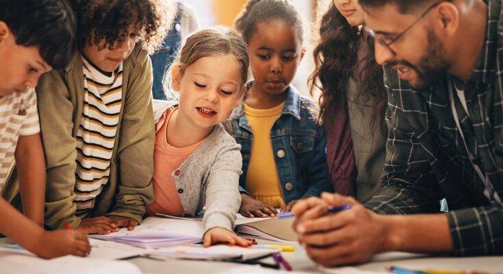 Elementary school students gather around a table with a young male teacher, who leans on to the table and the school work that is sitting on it