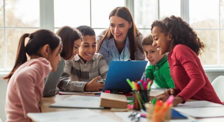 Young female teacher crouching down and smiling in front of a table of five elementary school age students.