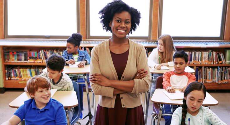 A young female Black teacher stands smiling at the camera with students sitting at desks behind her.