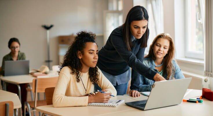 A teacher leans over a desk where two female high school students sit and review something on an open laptop.