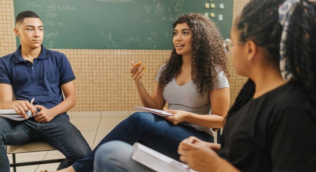 Group of college aged students sit in chairs while talking about a class assignment as a form of academic support. A young Black female student gestures and speaks to the group.