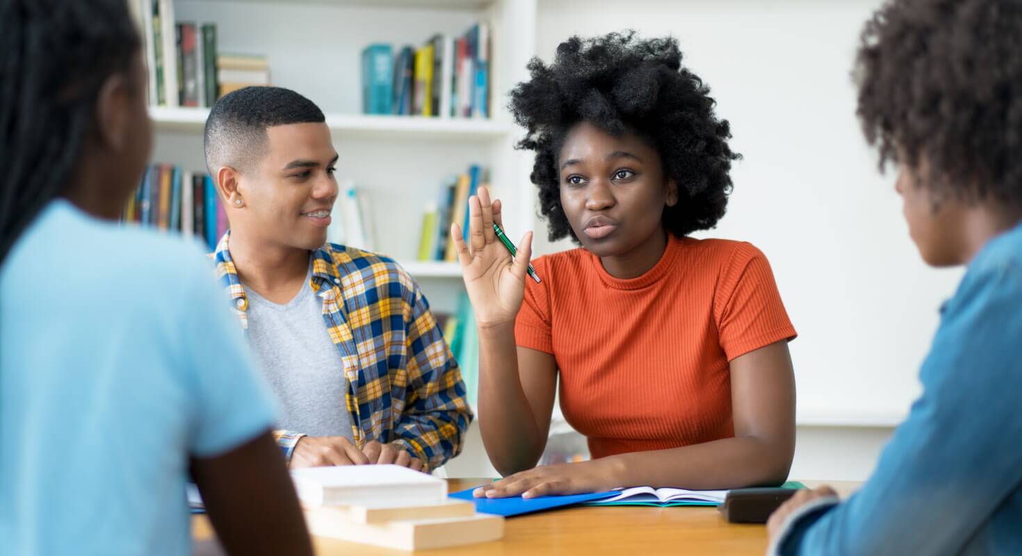 Group of college aged students sit in a peer study group to build academic support. A Black female student in an orange shirt gestures and speaks to the group.