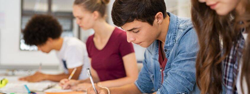 Group of college students studying in classroom writing notes during lesson. Focused guy and girls studying in college library sitting at desk. Group of multiethnic university students doing research sitting in a row.