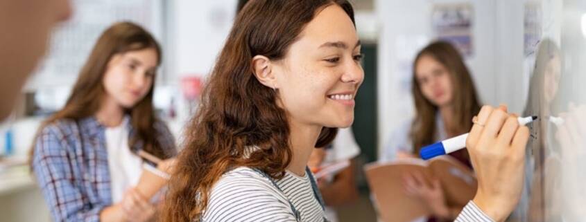 Female teenage high school student with long brown hair smiles as she writes on a white board. Other students look on in the background.