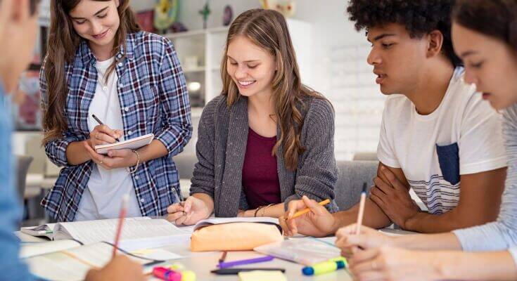 High school student sits at a table with several other students looking on . The students are taking notes with books and paper spread out on the table.