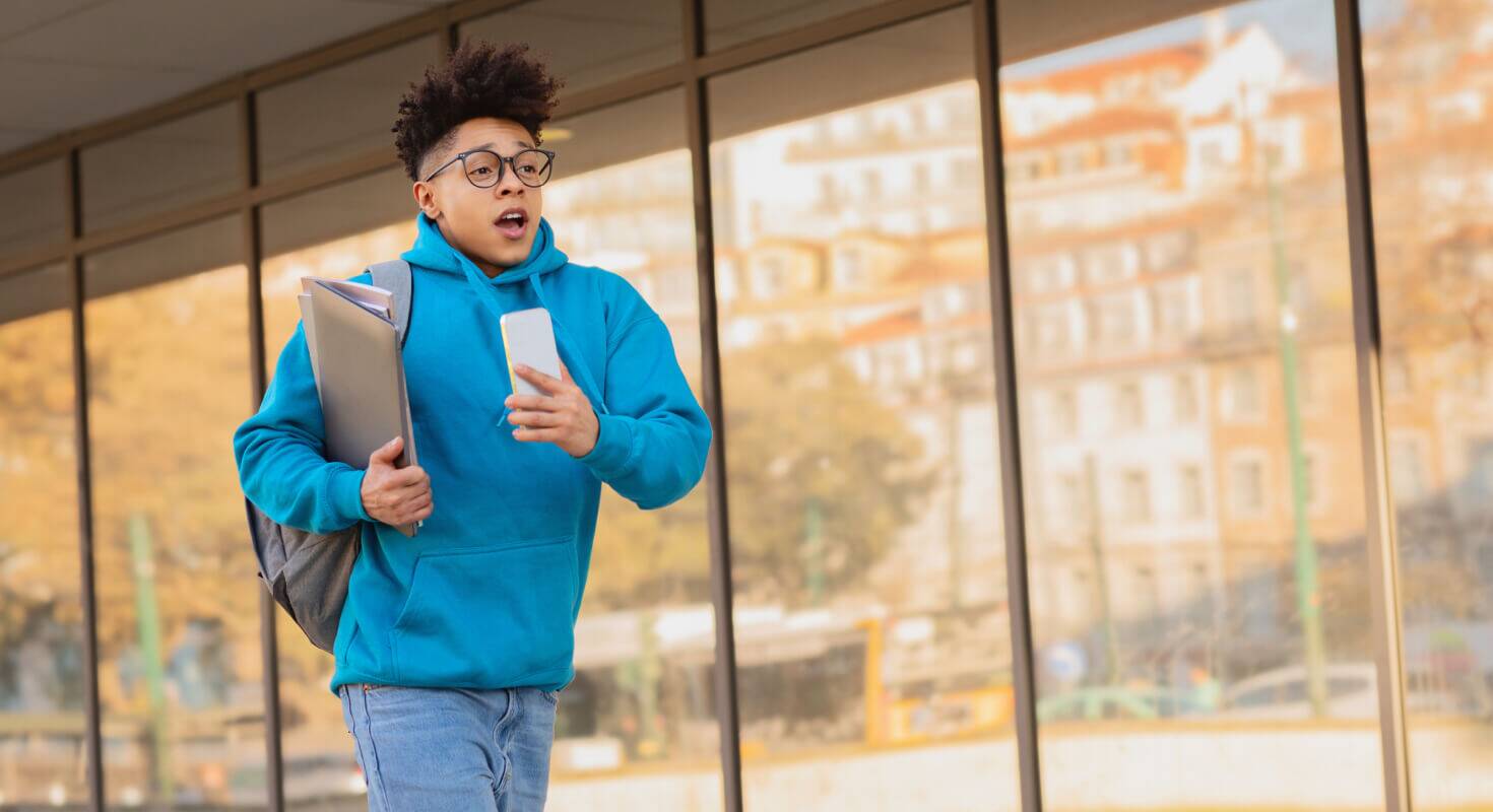 Middle school boy in blue sweatshirt and holding a backpack is running in front of the school as if he's late
