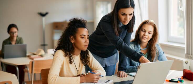 A teacher leans over a desk where two female high school students sit and review something on an open laptop.