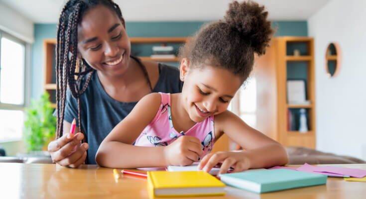Elementary school age child sits in front of her mom at the kitchen table with paper and crayons in front of them.