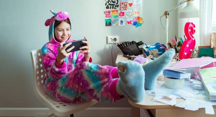 Adolescent student sits with her feet up on the desk and phone in her hands, ignoring her homework.