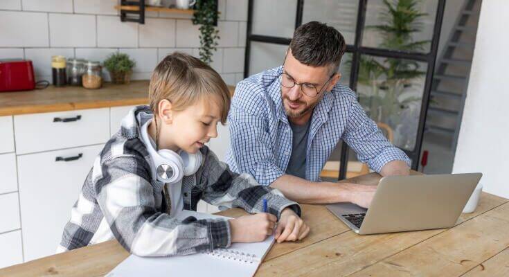 Older elementary school male student sits at the kitchen table with his dad, with the student writing on a piece of paper while the dad leans over and looks on.