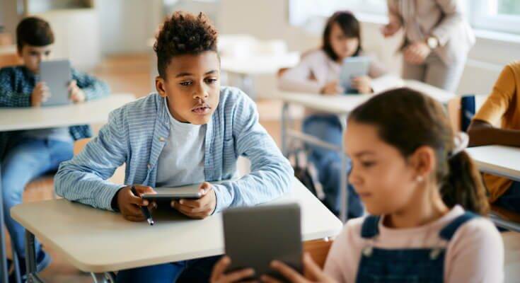 A male middle school student leans forward at his desk while holding a tablet, looking at the tablet of a female student sitting in front of him, as she points out something on the tablet.