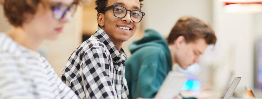 Cheerful excited smart black student in glasses sitting at table and looking at camera while using laptop in library