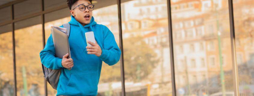 Middle school boy in blue sweatshirt and holding a backpack is running in front of the school as if he's late