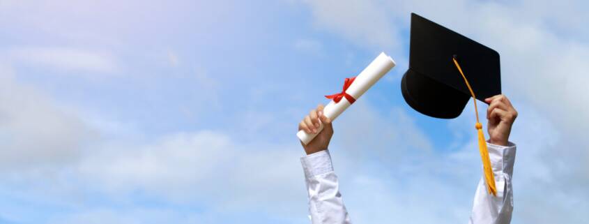 Man's arms raised against a blue sky holding a graduation cap and rolled diploma